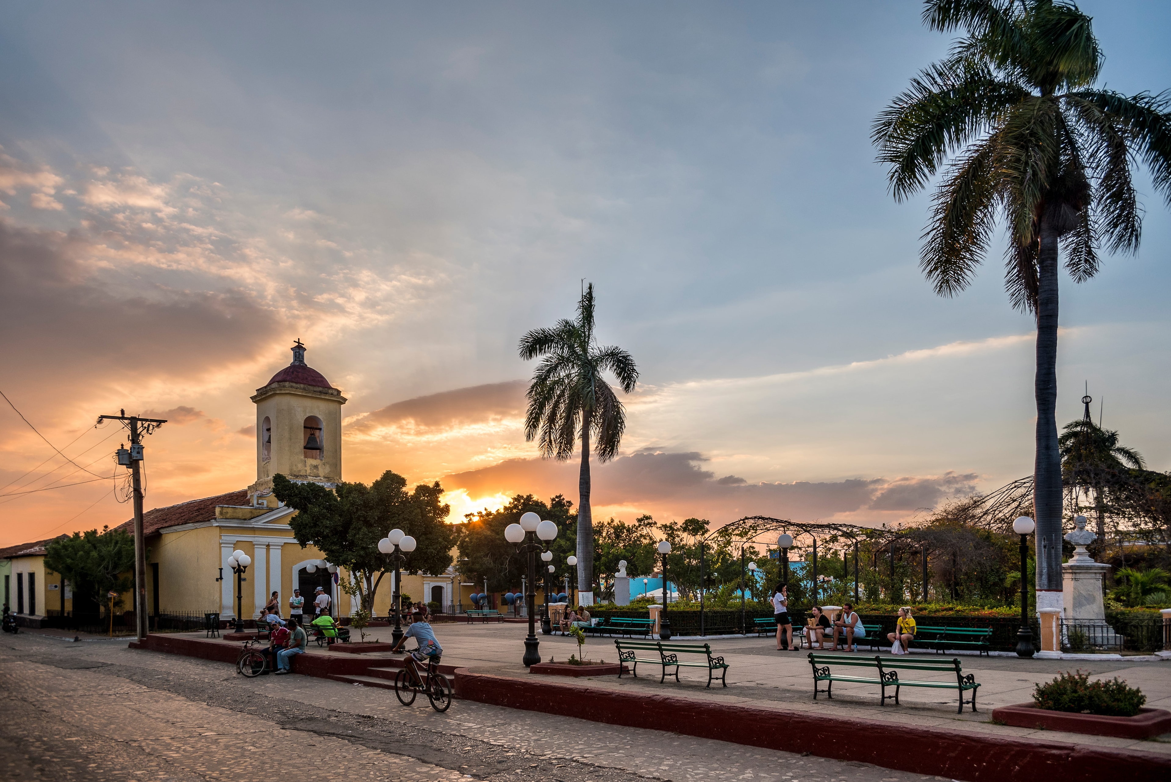 Trinidad Cuba town square at sunset with yellow church bell tower, royal palms, and locals on benches, Cuba