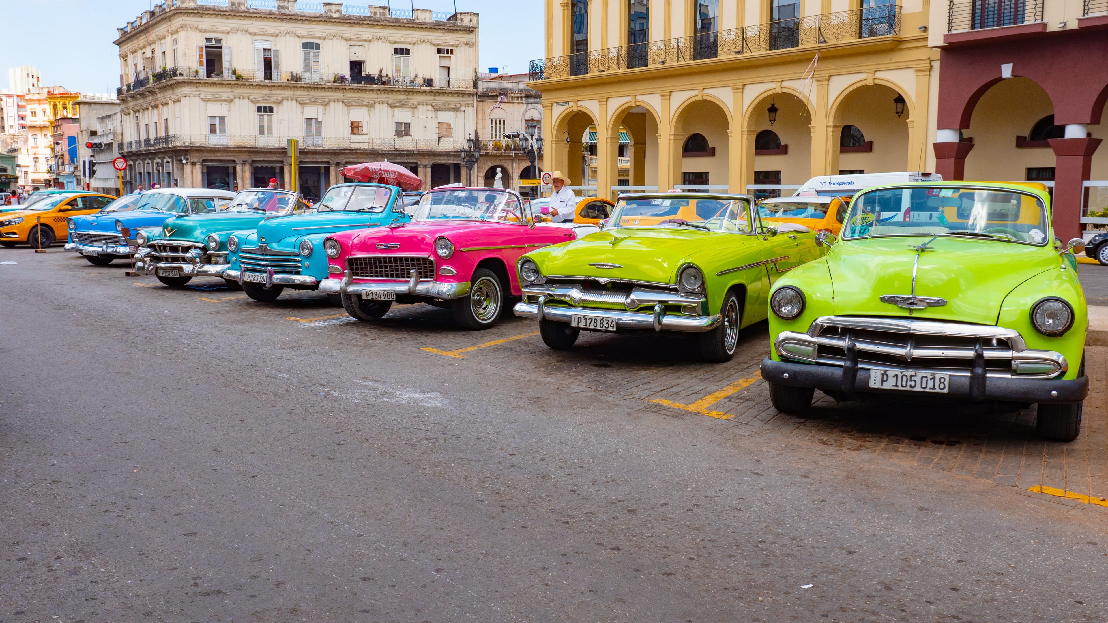 Row of colorful vintage convertibles — blue, teal, pink, and lime green — parked in front of colonial arcade, Havana Cuba