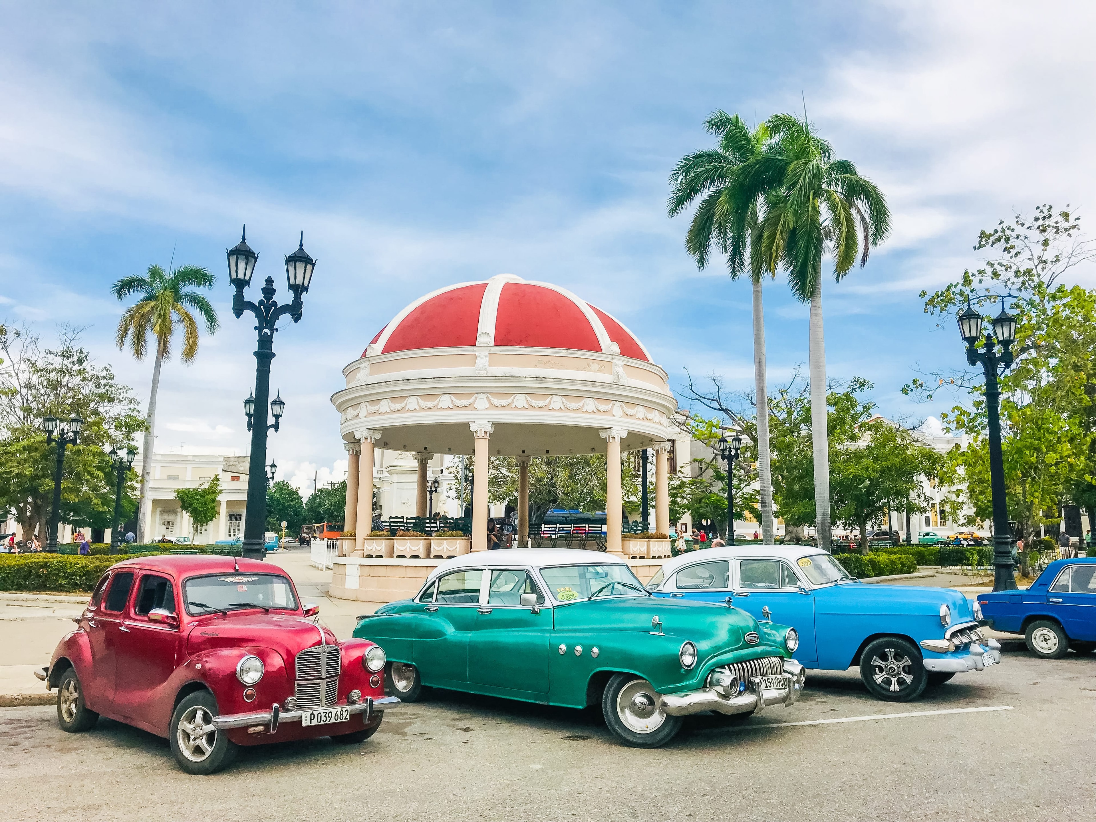 Red, teal, and blue vintage cars parked in front of red-domed gazebo in Cienfuegos Parque José Martí, Cuba