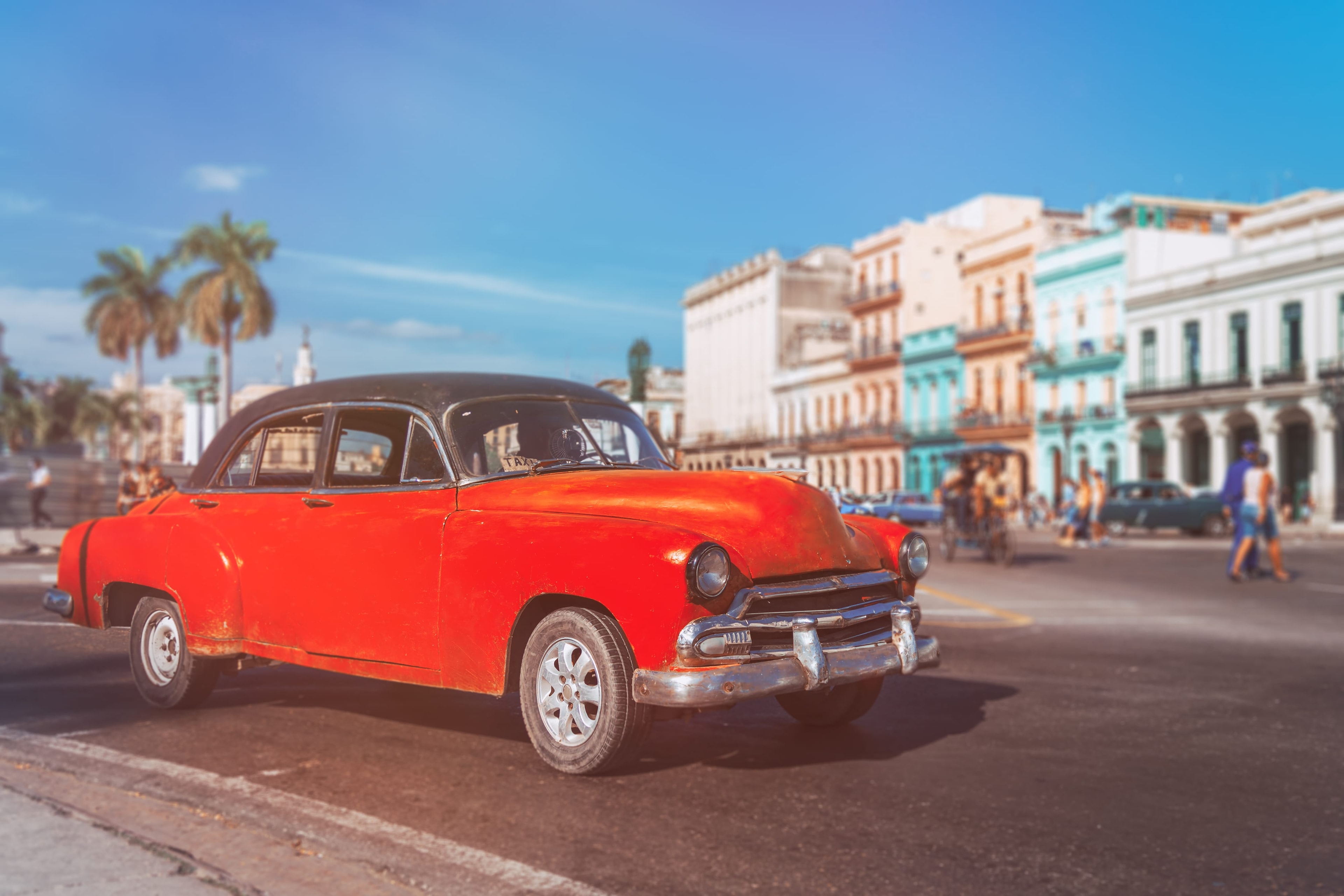 Red vintage car with black roof driving on Havana boulevard past pastel colonial buildings and palm trees, Cuba