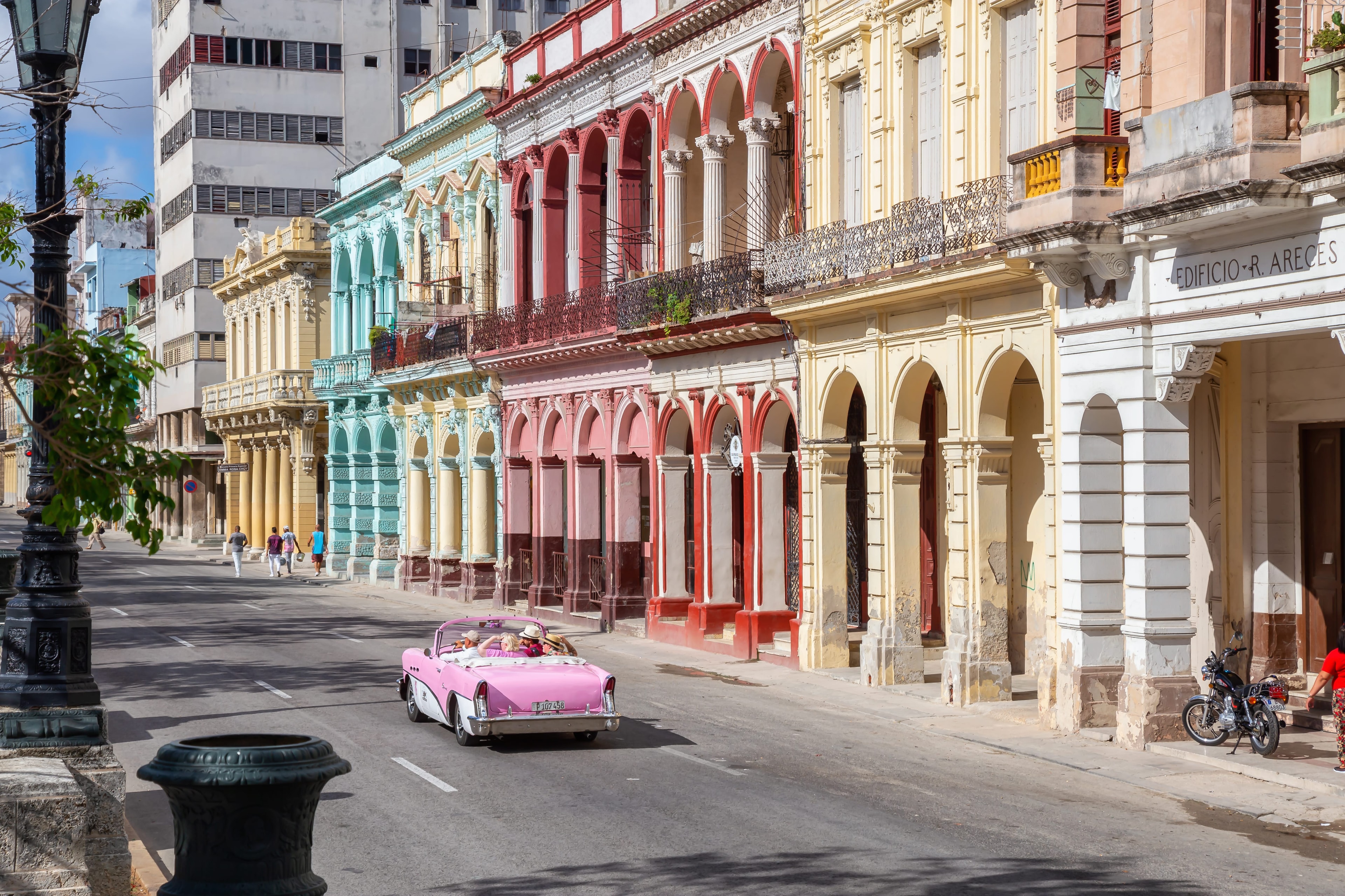 Pink vintage convertible cruising past row of colorful colonial arcaded facades — teal, crimson, and yellow — on Havana boulevard, Cuba