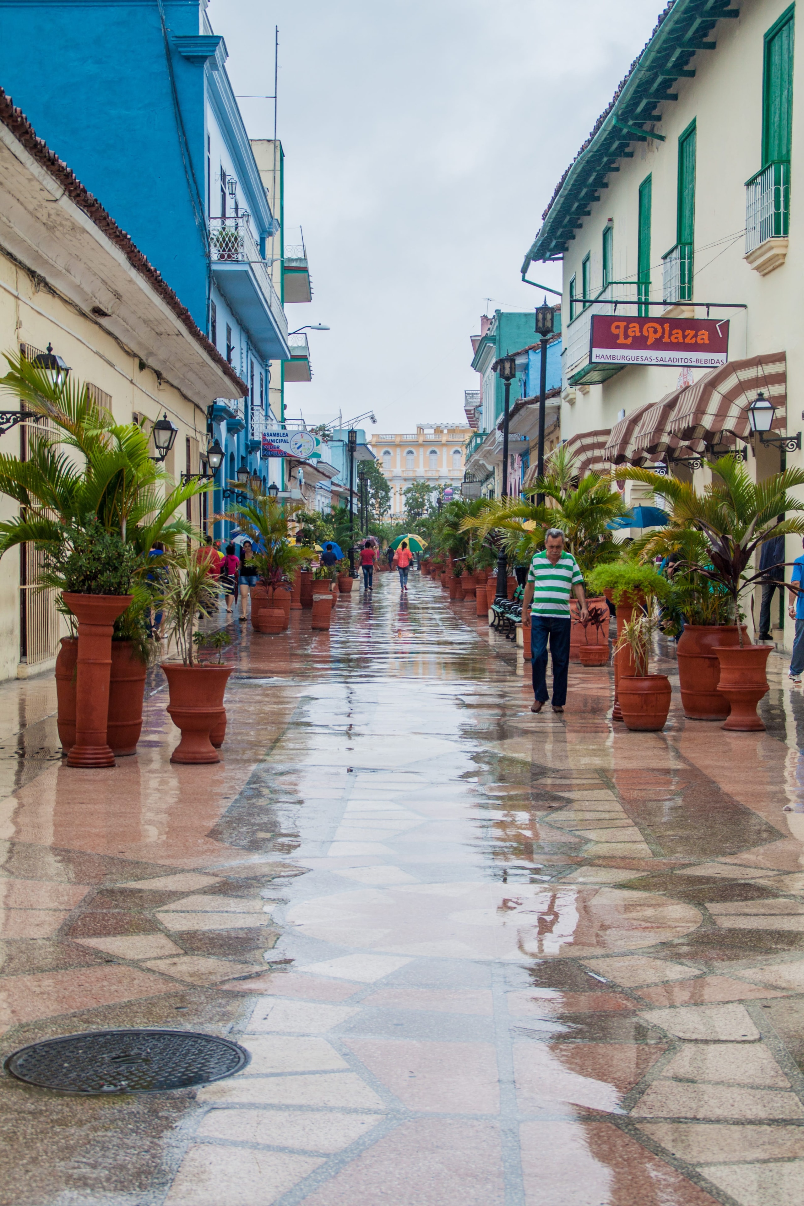 Rain-soaked pedestrian boulevard in Sancti Spíritus with terracotta planters, colorful buildings, and La Plaza sign reflecting in wet tiles, Cuba