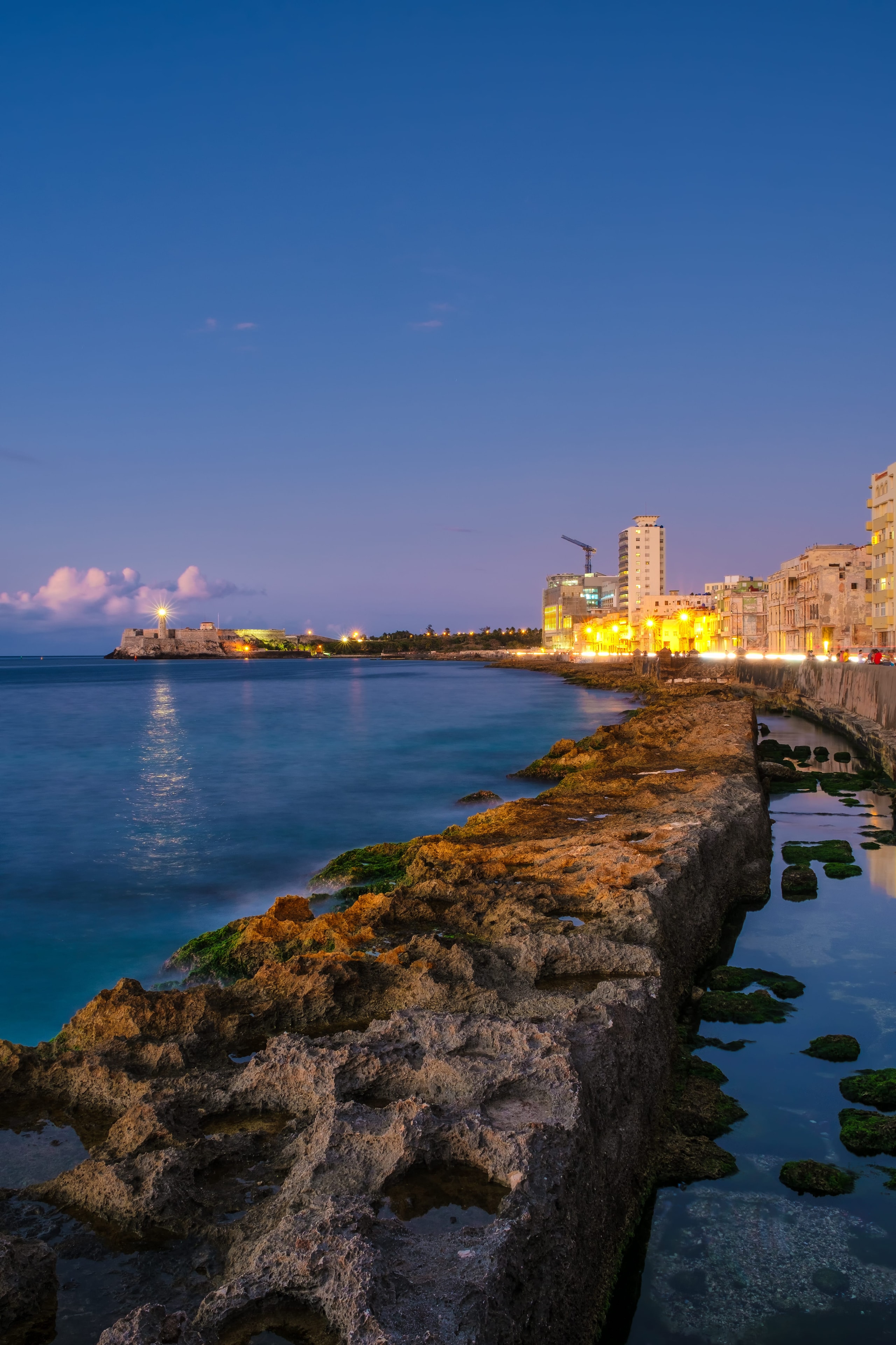 Havana Malecón seawall at blue hour with rocky shoreline, city lights, and Castillo del Morro lighthouse glowing across the bay, Cuba