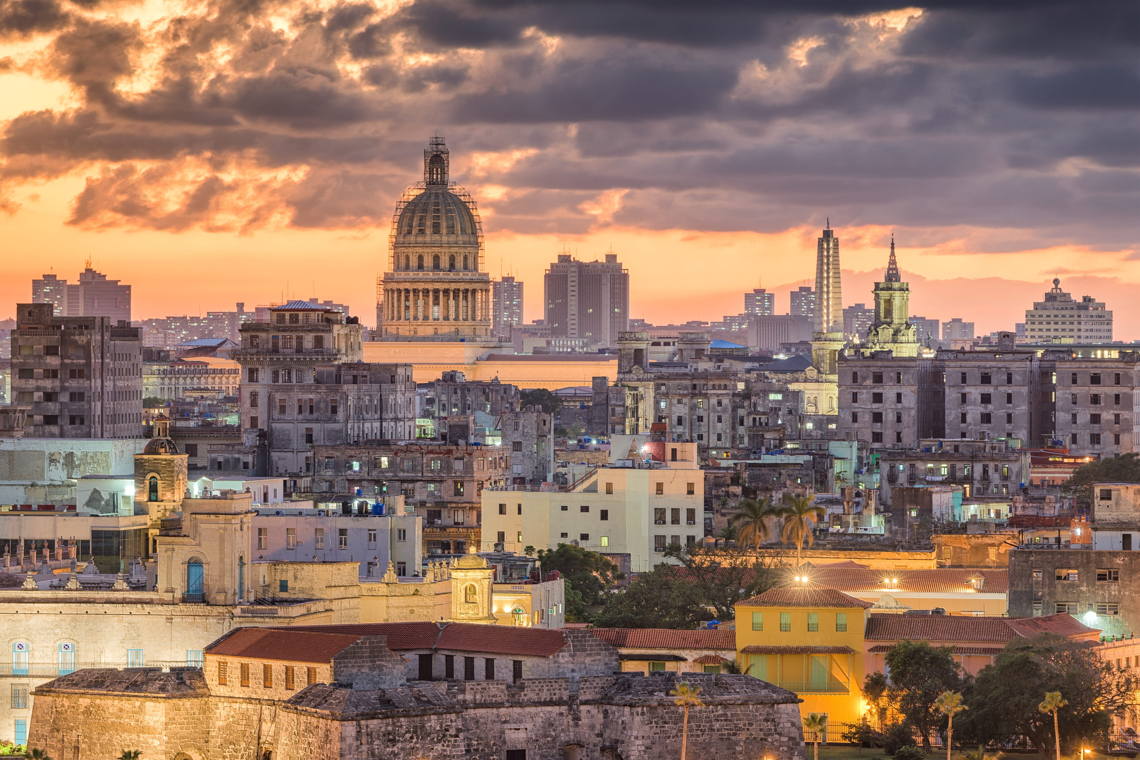 Havana skyline at dusk with El Capitolio dome under stormy orange sky, city lights beginning to glow across the rooftops, Cuba