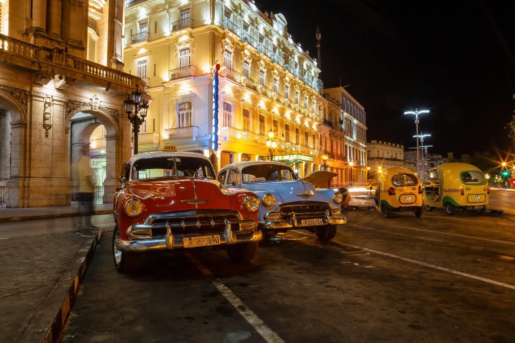 Vintage red and blue classic cars parked at night outside illuminated Havana hotel